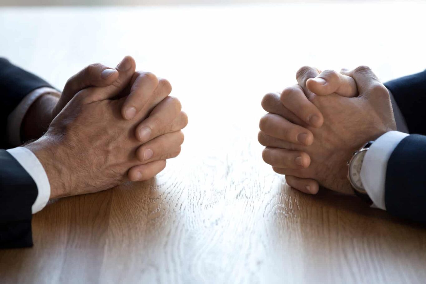 Dois homens de mãos cruzadas sentam-se frente a frente num cenário de confronto.